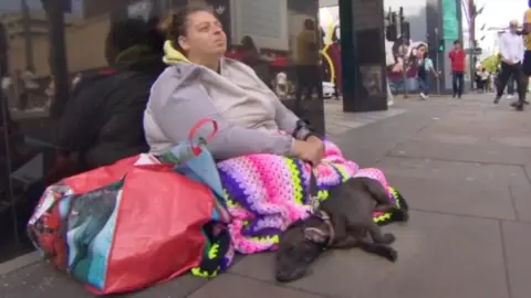 Adele, a homeless woman living in Manchester, sits outside a high street department store with a colorful blanket over her knees, her labrador Belle lying at her feet.