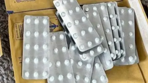 Several silver blister packs of white pills, lying on top of brown manilla envelopes