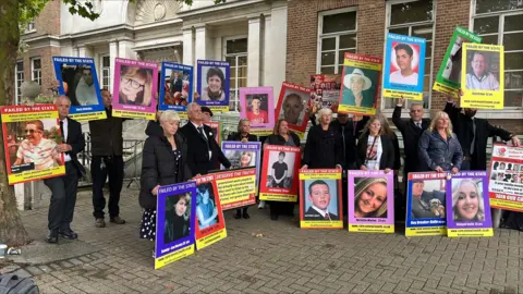 RICHARD KNIGHTS/BBC A large group of grieving family members stand outside the Civic Center in Chelmsford on the first day of The Lampard Inquiry holding large posters of their loved ones with captions such as "failed by the state". They are wearing coats and black clothes.