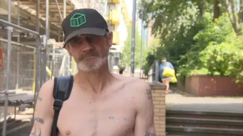 BBC Ricky, with a gray beard and wearing a black baseball cap, stands near the spot where one of his friends died of a drug overdose. There are tattoos on his bare chest to honor friends he has lost