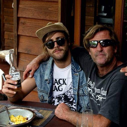 Two men in sunglasses sit at a table with their arms around each other.